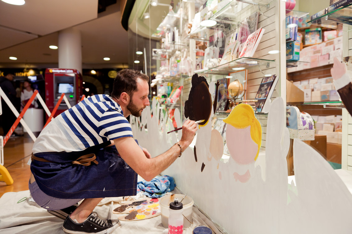 Matt squatting in front of a 5m mural concentrating on painting a small child character